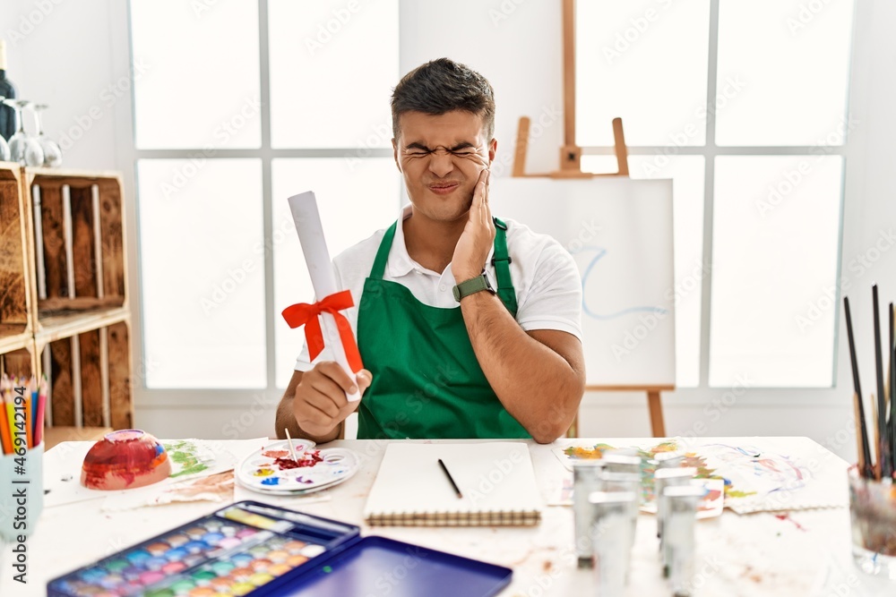 Young hispanic man at art studio holding degree touching mouth with hand with painful expression because of toothache or dental illness on teeth. dentist concept.