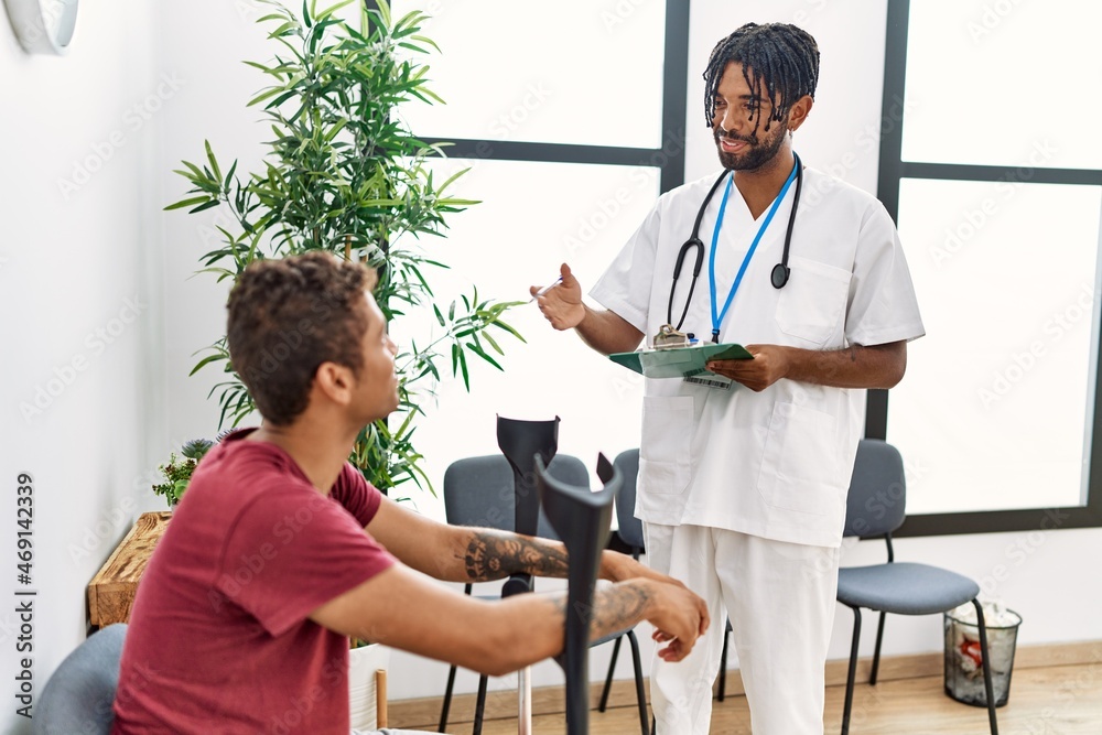 Fototapeta premium Two men physiptherapist and patient having medical consultation using crutches at hospital waiting room