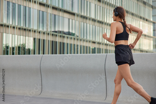 Active athletic woman with pigtail runs outdoors has slim body and muscular legs dressed in sportswear listens music in headphones being health conscious joggs at sidewalk demonstrates endurance