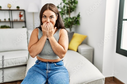 Young brunette woman sitting on the sofa at home laughing and embarrassed giggle covering mouth with hands, gossip and scandal concept