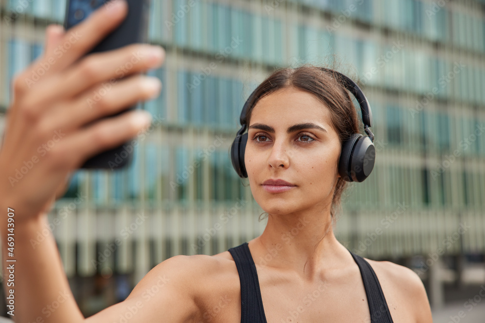 Serious athletic woman uses phone for online call or taking selfie on front camera during fitness workout has confident expression spends free time in urban place poses agains blurred background