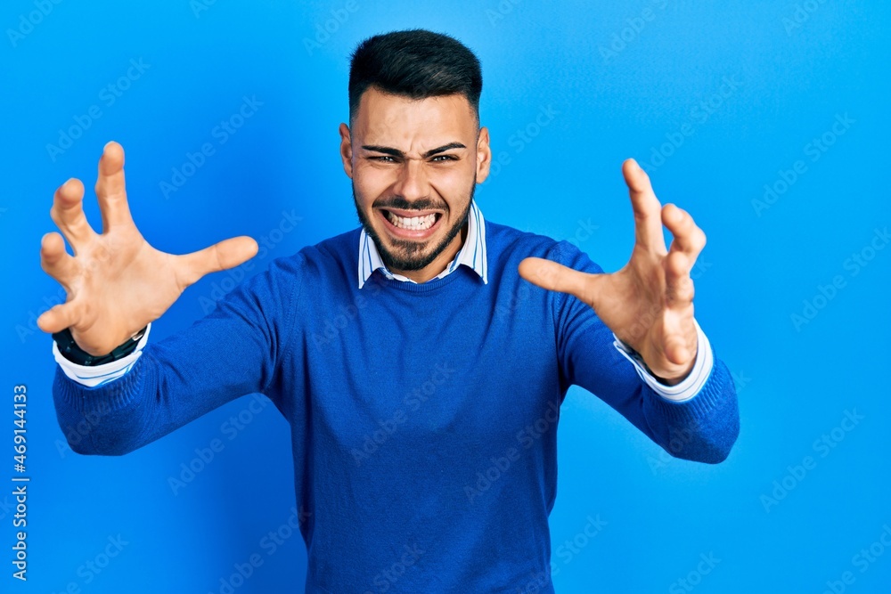 Young hispanic man with beard wearing casual blue sweater shouting frustrated with rage, hands trying to strangle, yelling mad Young hispanic man with beard wearing casual blue sweater shouting frustrated with rage, hands trying to strangle, yelling mad