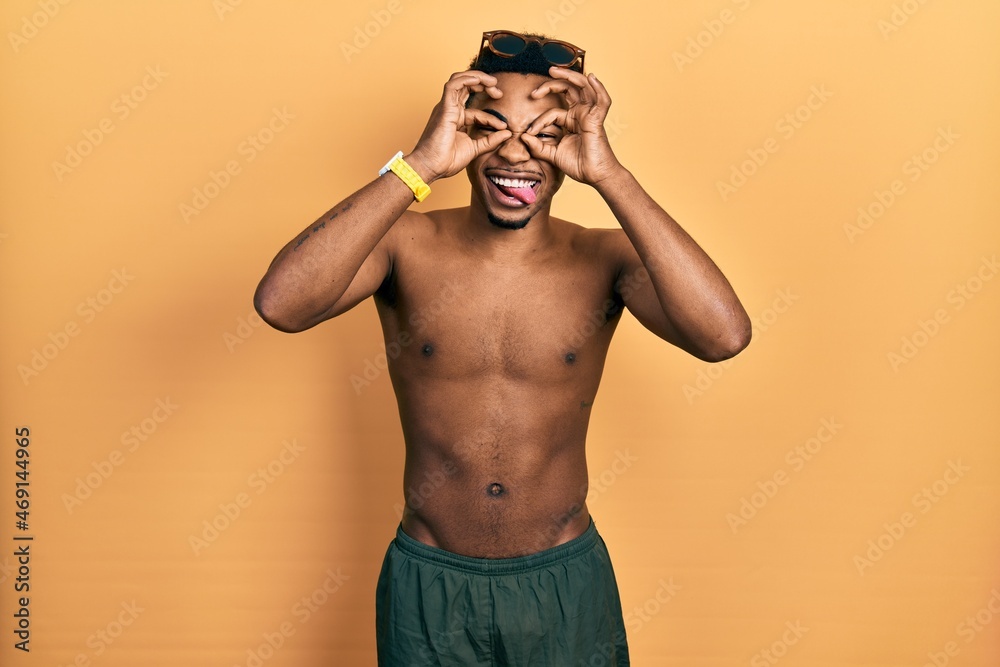 Fototapeta premium Young african american man wearing swimwear and swimmer glasses doing ok gesture like binoculars sticking tongue out, eyes looking through fingers. crazy expression.