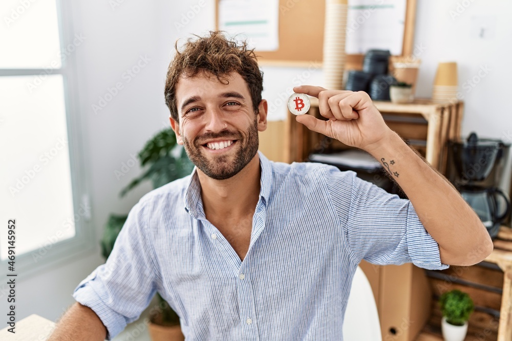 Young hispanic businessman smiling happy holding bitcoin at the office. Young hispanic businessman smiling happy holding bitcoin at the office.