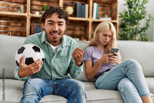 Man smiling happy watching soccer match and girlfriend boring using smartphone at home.