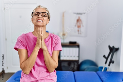 Middle age blonde woman at pain recovery clinic begging and praying with hands together with hope expression on face very emotional and worried. begging.