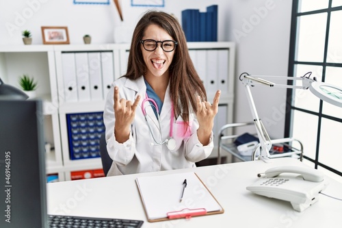 Young doctor woman wearing doctor uniform and stethoscope at the clinic shouting with crazy expression doing rock symbol with hands up. music star. heavy concept.
