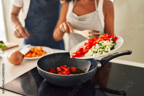 Middle age hispanic couple pouring food on frying pan at kitchen