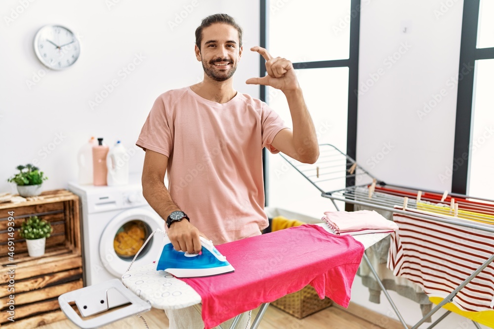 Obraz premium Young hispanic man ironing clothes at home smiling and confident gesturing with hand doing small size sign with fingers looking and the camera. measure concept.