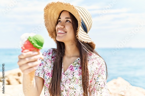 Young latin girl wearing summer hat eating ice cream sitting on the rock at the beach.