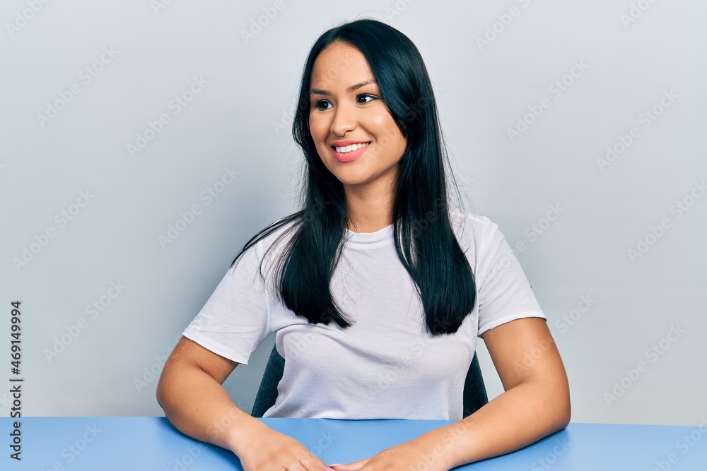Beautiful hispanic woman with nose piercing wearing casual white t shirt sitting on the table looking to side, relax profile pose with natural face and confident smile.