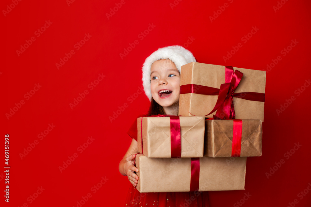 Merry Christmas and happy holidays. Portrait of a pending happy girl in a Santa cap with gifts boxes on a red background. Kids, children. A place for text. Merry Christmas and happy holidays. Portrait of a pending happy girl in a Santa cap with gifts boxes on a red background. Kids, children. A place for text.