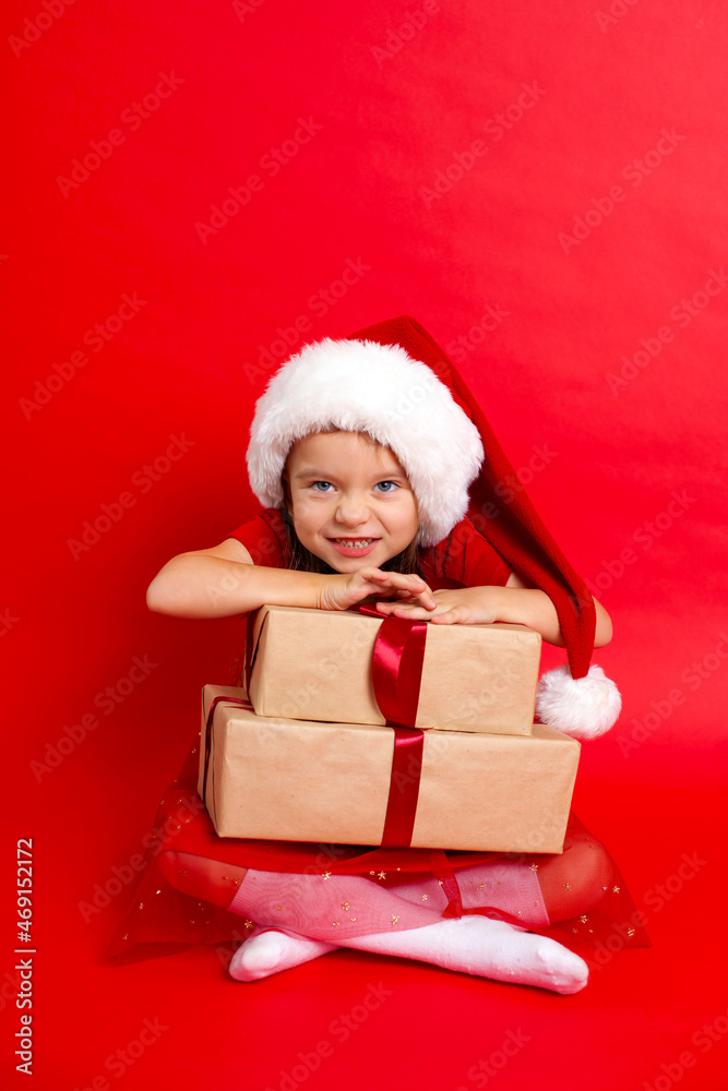 Merry Christmas and happy holidays. Portrait of a pending happy girl in a Santa cap with gifts boxes on a red background. Kids, children. A place for text. Merry Christmas and happy holidays. Portrait of a pending happy girl in a Santa cap with gifts boxes on a red background. Kids, children. A place for text.