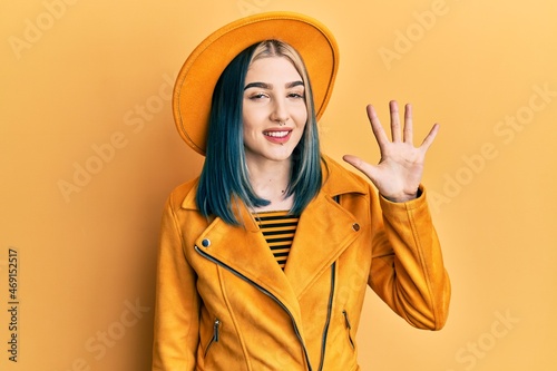 Young modern girl wearing yellow hat and leather jacket showing and pointing up with fingers number five while smiling confident and happy. © Krakenimages.com