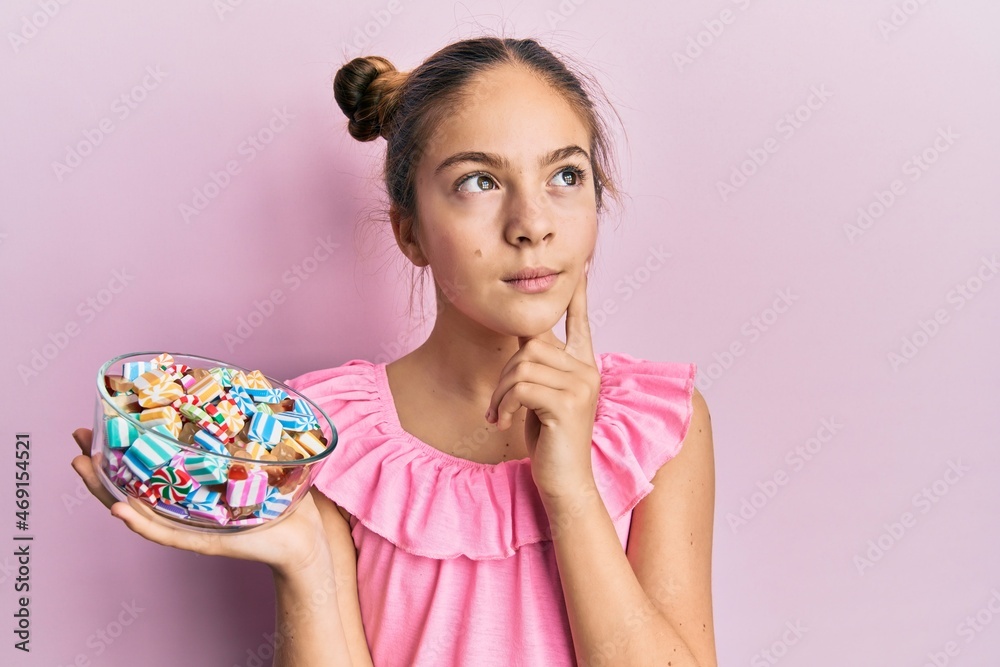 Beautiful brunette little girl holding jar full of sugar candies serious face thinking about question with hand on chin, thoughtful about confusing idea Beautiful brunette little girl holding jar full of sugar candies serious face thinking about question with hand on chin, thoughtful about confusing idea