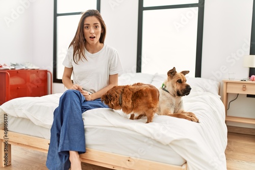Young brunette woman sitting on the bed with two dogs scared and amazed with open mouth for surprise, disbelief face