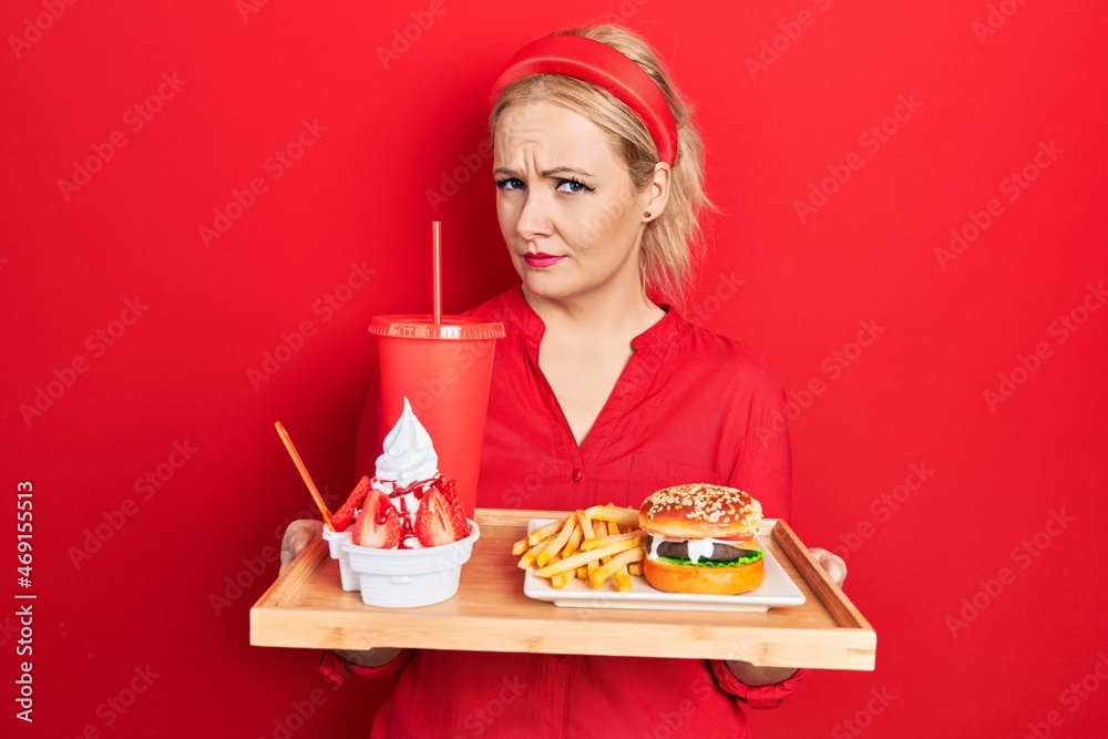 Young blonde woman eating a tasty classic burger with fries and soda clueless and confused expression. doubt concept. Young blonde woman eating a tasty classic burger with fries and soda clueless and confused expression. doubt concept.