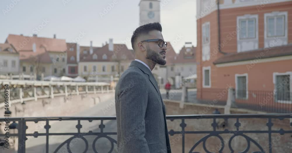 side view of elegant man wearing suit and long coat, walking in a medieval city, looking away, smiling and military saluting side view of elegant man wearing suit and long coat, walking in a medieval city, looking away, smiling and military saluting