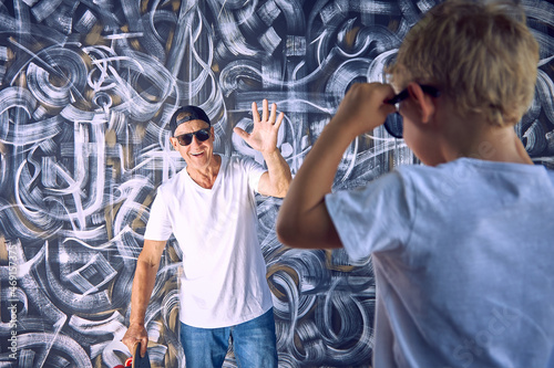 Slika na platnu Cool sports grandfather with a skateboard in his hand waves to a surprised grandson in sunglasses