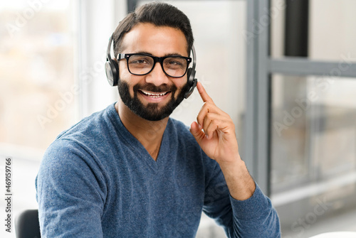 Headshot of young Indian male wearing handsfree headset and stylish glasses, in casual wear in the office, taking call and talking with customers or colleagues. Call center, help line, support service