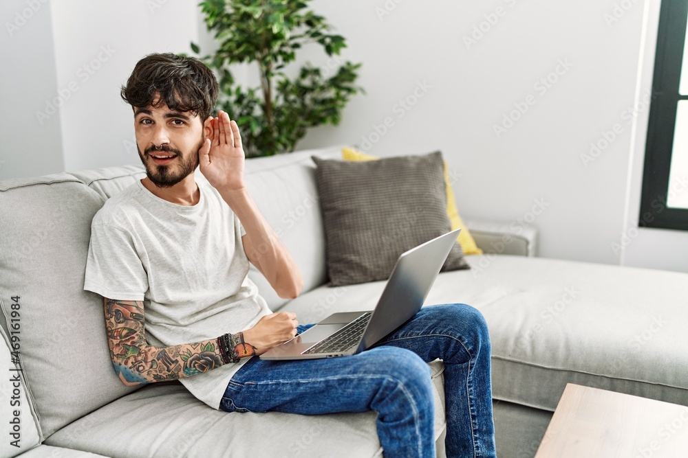 Hispanic man with beard sitting on the sofa smiling with hand over ear listening an hearing to rumor or gossip. deafness concept.