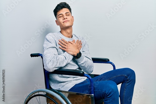 Young hispanic man sitting on wheelchair smiling with hands on chest with closed eyes and grateful gesture on face. health concept.