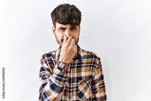 Hispanic man with beard standing over isolated background smelling something stinky and disgusting, intolerable smell, holding breath with fingers on nose. bad smell