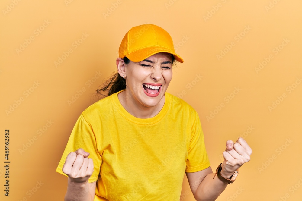 Young hispanic woman wearing delivery uniform and cap very happy and excited doing winner gesture with arms raised, smiling and screaming for success. celebration concept.