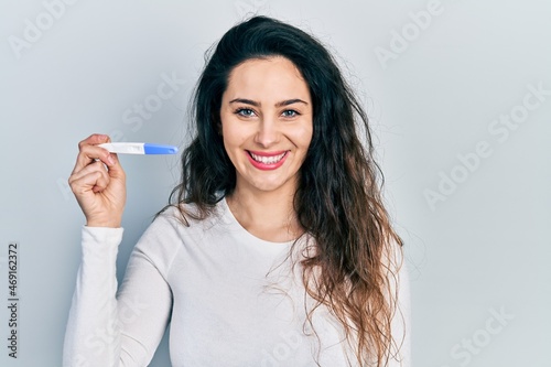 Young hispanic woman holding thermometer looking positive and happy standing and smiling with a confident smile showing teeth
