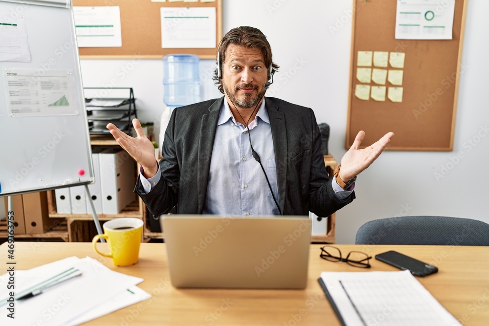 Handsome middle age man wearing call center agent headset at the office clueless and confused expression with arms and hands raised. doubt concept. Handsome middle age man wearing call center agent headset at the office clueless and confused expression with arms and hands raised. doubt concept.