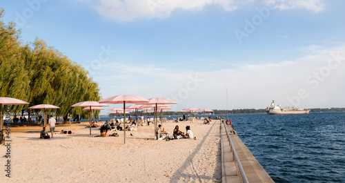 Toronto, Canada - 08 29 2021: Torontonians in downtown Toronto having rest under pink umblrellas of the Sugar Beach bestde blue waters of lake Ontario with a cargo ship