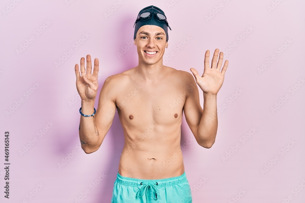Young hispanic man wearing swimwear and swimmer glasses showing and pointing up with fingers number nine while smiling confident and happy.