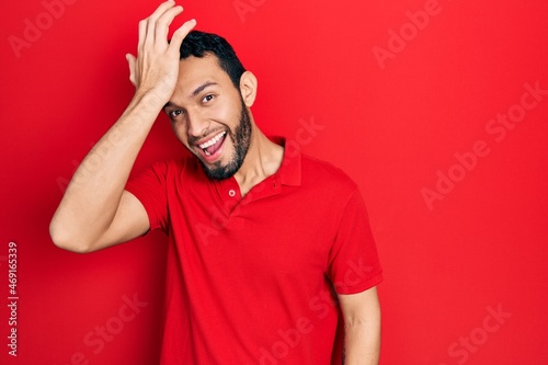 Hispanic man with beard wearing casual red t shirt surprised with hand on head for mistake, remember error. forgot, bad memory concept.