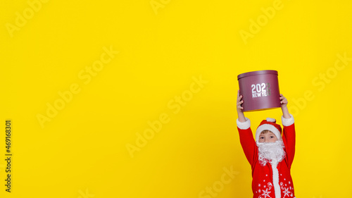 A small Caucasian child in Santa Claus clothes and with an artificial white beard high above his head is holding a large round gift box with the words New Year 2022, selective focus © Aleksandr Uglov