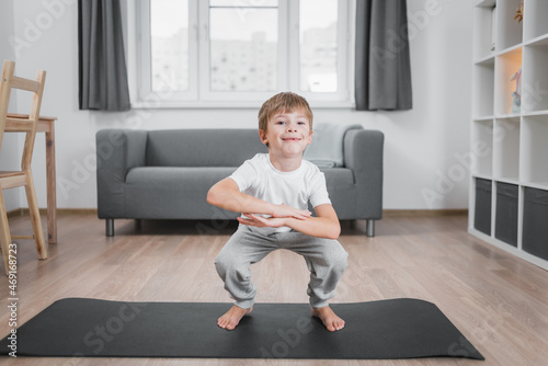 Boy-child doing classic squats at home on the floor on a sports mat, wearing a white t-shirt and gray pants.