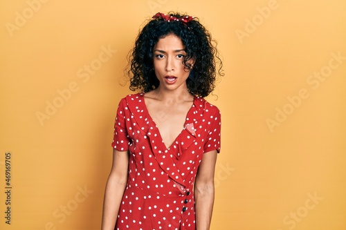 Young latin girl wearing summer dress in shock face, looking skeptical and sarcastic, surprised with open mouth