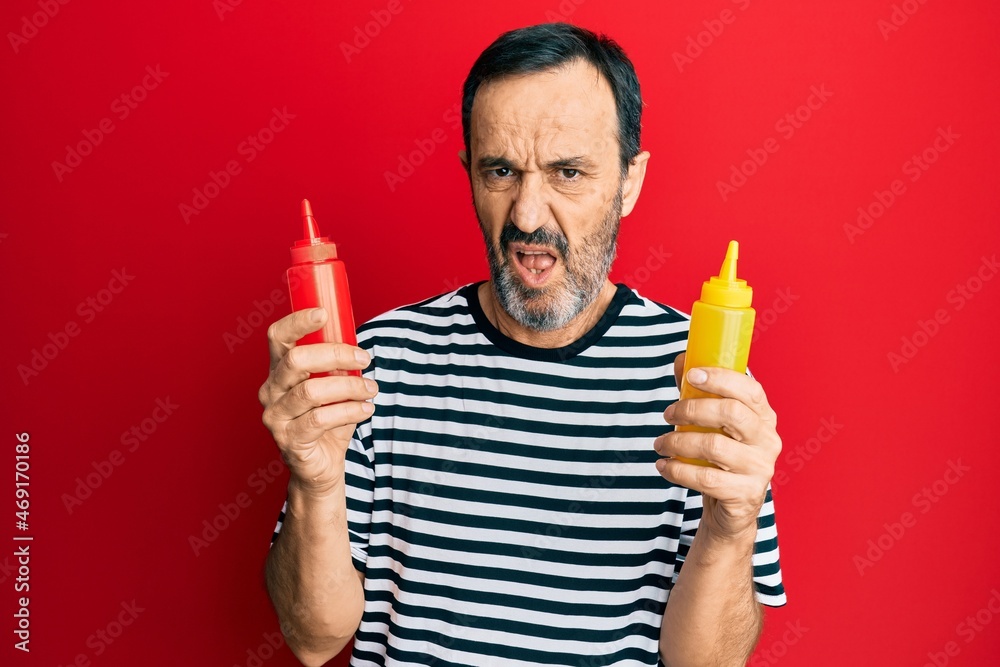 Middle age hispanic man holding ketchup and mustard bottle clueless and confused expression. doubt concept. Middle age hispanic man holding ketchup and mustard bottle clueless and confused expression. doubt concept.