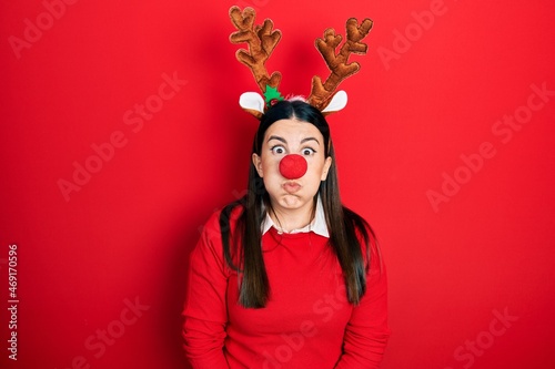 Young hispanic woman wearing deer christmas hat and red nose puffing cheeks with funny face. mouth inflated with air, crazy expression.