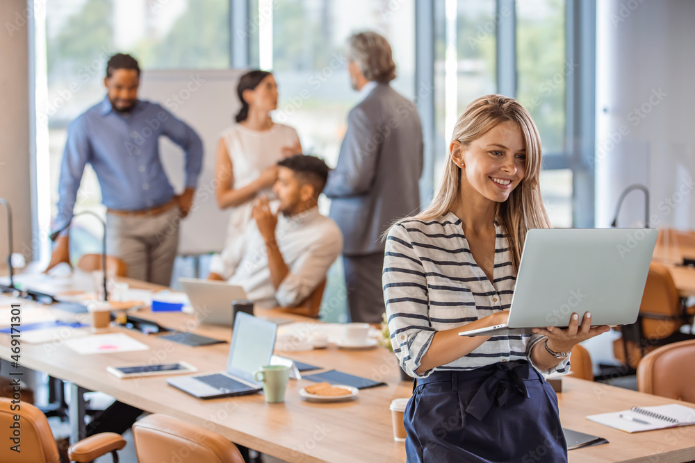 Woman manager holding laptop and standing in modern office. Businesswoman in casual wear holding laptop smiling and looking to camera while her colleagues communicating in the background Woman manager holding laptop and standing in modern office. Businesswoman in casual wear holding laptop smiling and looking to camera while her colleagues communicating in the background