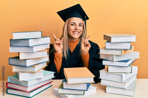 Young caucasian woman wearing graduation ceremony robe sitting on the table smiling confident pointing with fingers to different directions. copy space for advertisement