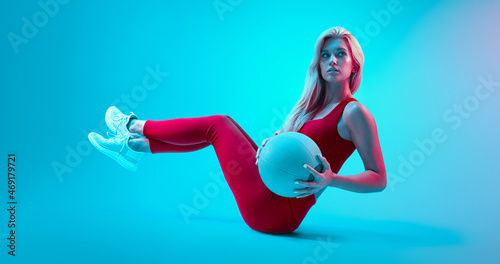 Fitness woman working out with fitness ball on bright neon cyan background. Neon blue light shot of strong muscular young woman holding ball