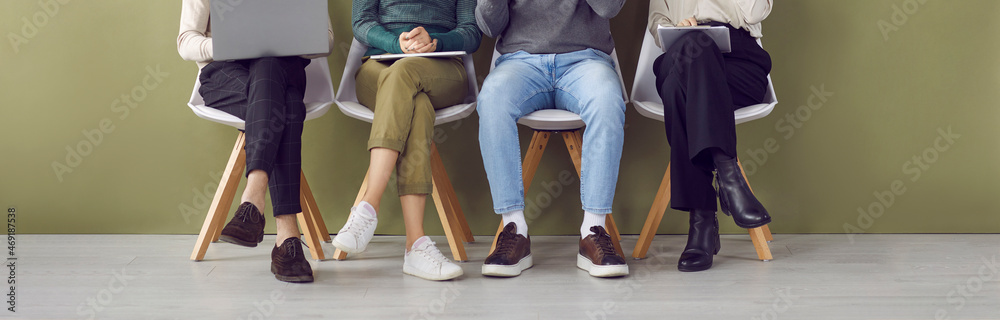 Legs of four job applicants sitting on chairs in hallway waiting in line for interview. Unknown candidates for jobs in casual clothes and shoes are sitting with documents and gadgets. Hiring concept. - obrazy, fototapety, plakaty 