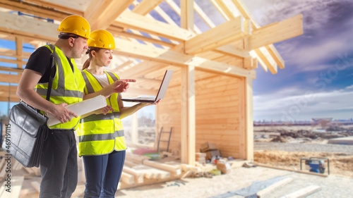 Builders with laptop. Man and woman are working as builders. Work in construction company. Timber house frame in background. Career in architectural bureau. Constructors-designers.