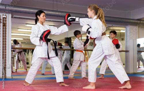 Young girls and boys in boxing gloves exercising jabs during group karate training. Trainer with focus mitts teaching girl in foreground.