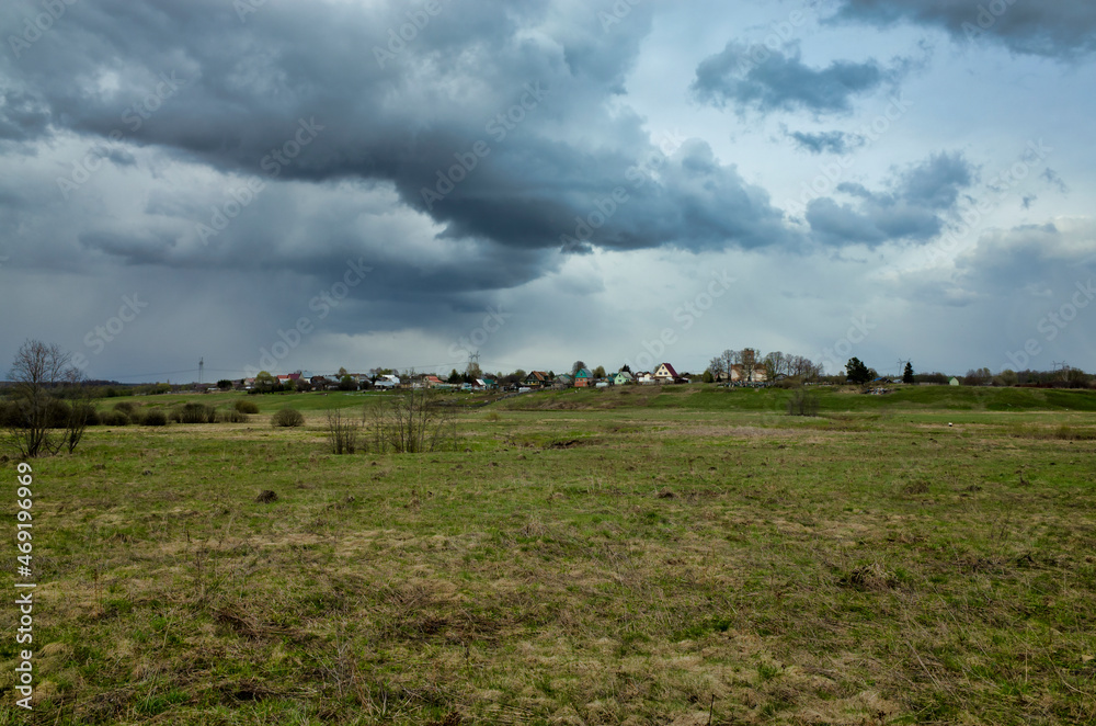 Obraz premium Spring meadow overlooking the village. The sky is covered with rain clouds. Nature of the Vladimir region, Russia