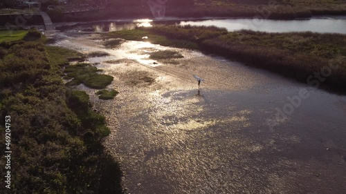 Cinematic drone shot of surfer man taking surfboard over head after finishing surfing on Vicente Lopez River,Buenos Aires - Beautiful sunset light reflecting in water ponds of river shore photo