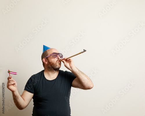 Portrait of Caucasian man celebrating joyfully. He has birthday hat, rattle, bugle and funny cotillion glasses. Neutral background. Horizontal image with copy space for text.