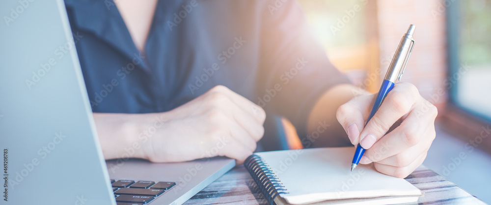 Woman hand writing on a notepad with a pen and use a laptop computer in the office.