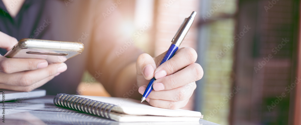Woman hand writing on a notepad with a pen and is using a mobile phone in the office.