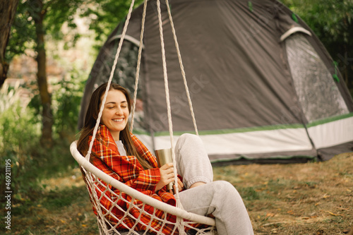 Beautiful Girl wrapped in red plaid drinking tea in a cozy hanging chair outdoors. Girl holding mug of hot tea. Adventure travel, camping and hiking in nature.